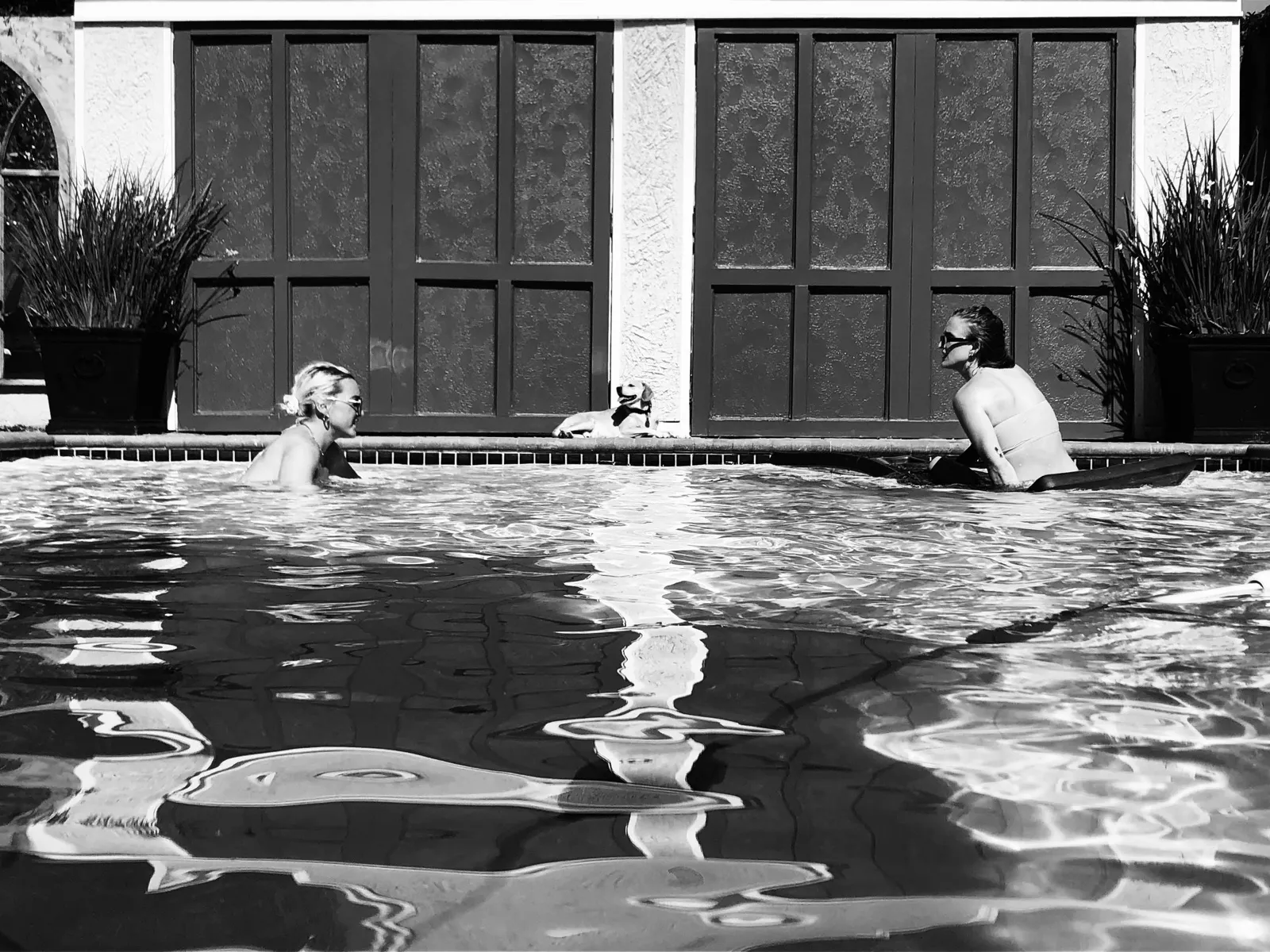Untitled (The Pool), black and white photograph by Mathieu: two women and a dog at a courtyard pool in flat afternoon light