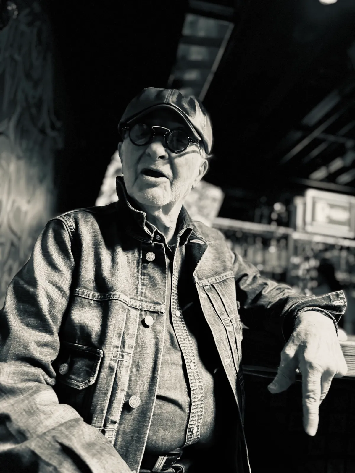 Untitled (The Bar), black and white photograph by Mathieu: an older man in a denim jacket, round sunglasses, and leather cap inside a bar