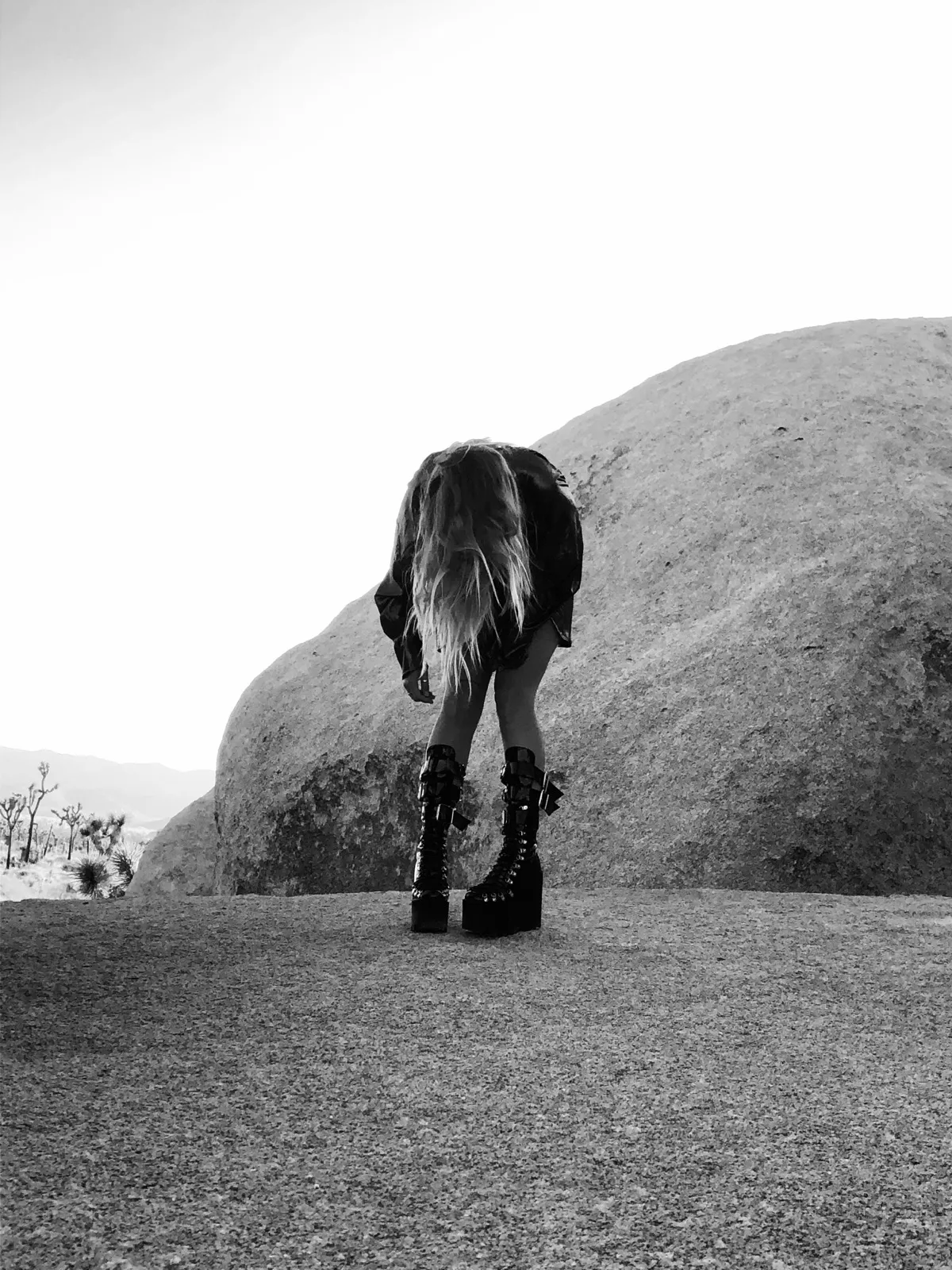 Untitled (Desert), black and white photograph by Mathieu: a figure bent over on desert rock under a white sky