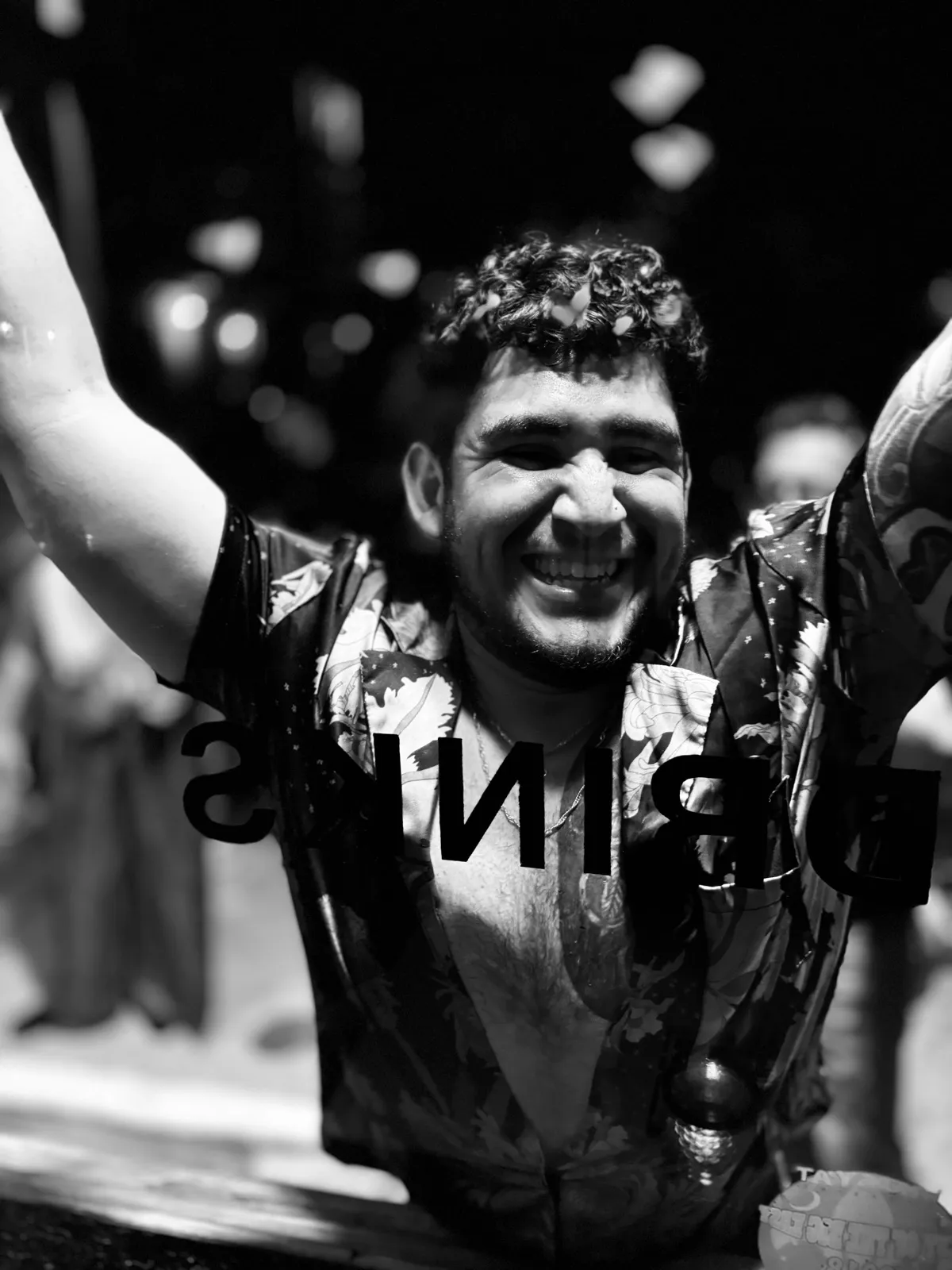 Untitled (Drinks), black and white photograph by Mathieu: a man smiling with raised arms behind a window lettered DRINKS