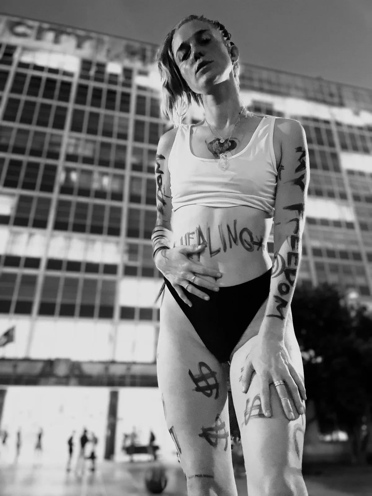 Untitled (Healing), black and white photograph by Mathieu: a woman with handwritten words on her skin standing before a city building