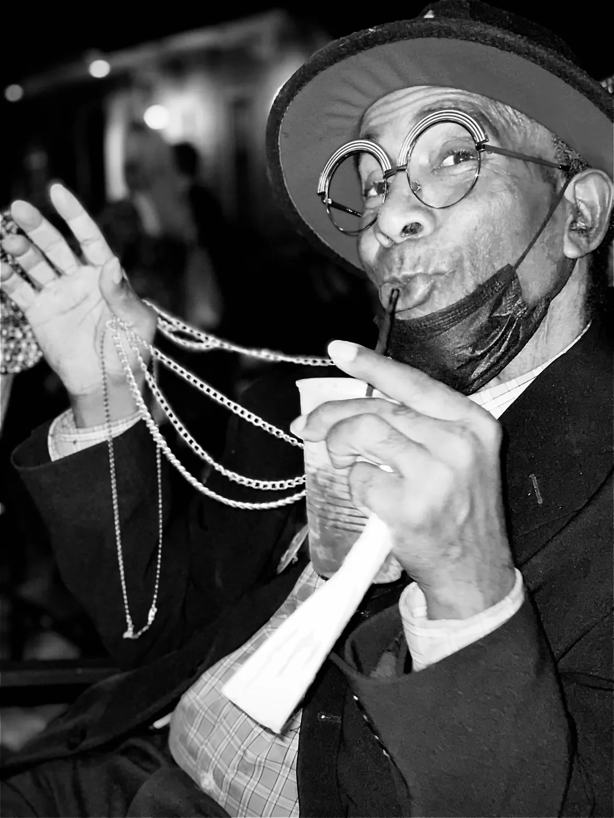 Untitled (Round Glasses), black and white photograph by Mathieu: a man in a hat and round glasses sipping through a straw, beads in hand