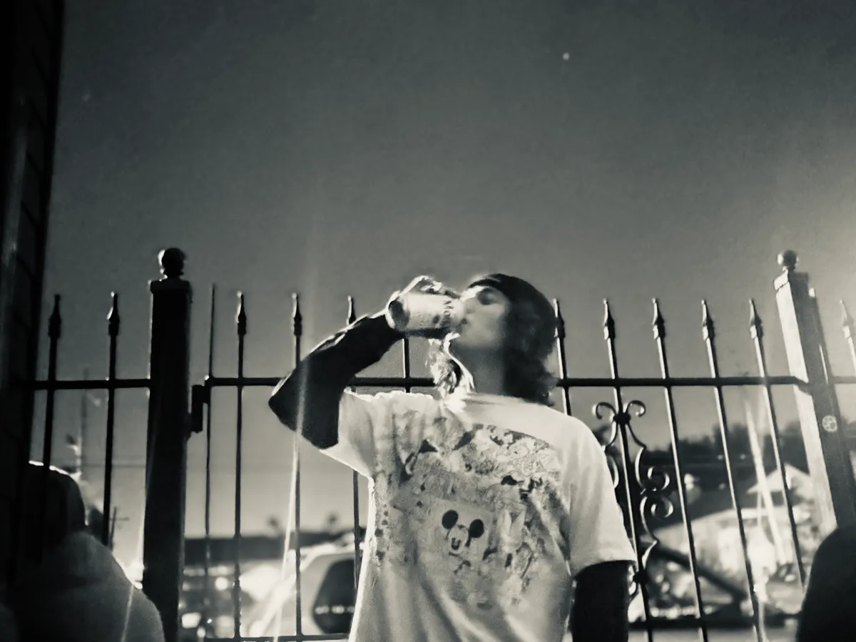Untitled (The Fence), black and white photograph by Mathieu: a figure drinking from a can against an iron fence and night sky