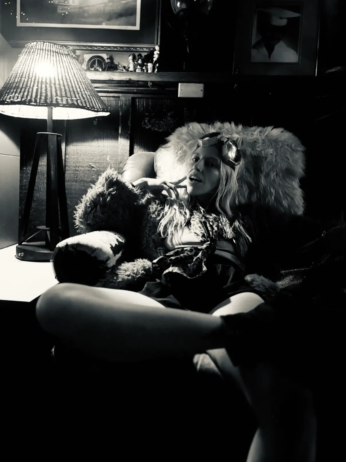 Untitled (The Lamp), black and white photograph by Mathieu: a woman in fur and goggles reclining beside a lit lamp in a dark interior